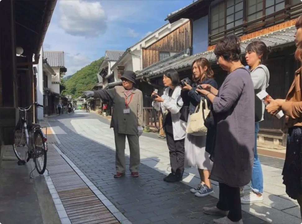 역사적인 창고와 마치야가 늘어선 다케하라 거리 보존지구의 거리에서, 가이드의 설명을 들으며 DSLR 카메라 등으로 열심히 촬영을 즐기는 관광객 그룹.