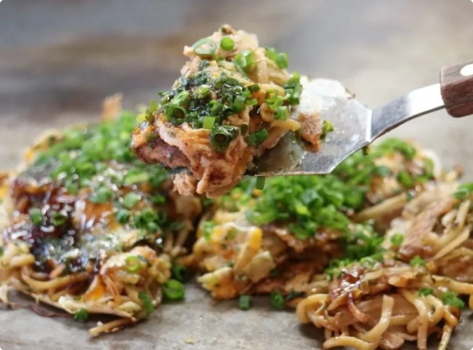 Hiroshima-style okonomiyaki topped generously with green onions, being lifted with a spatula on a hot griddle