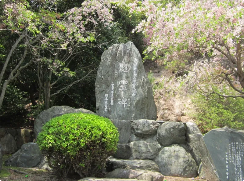 만개한 벚꽃 나무 아래 정적 속에 서 있는, 커다란 자연석으로 만들어진 '위령비'라고 새겨진 석비. 앞쪽에는 잘 가꾸어진 조경이 있으며 주변은 풍부한 녹음으로 둘러싸여 있다.