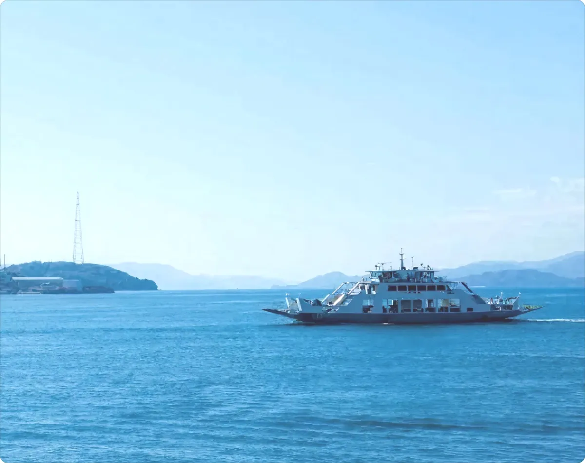 맑고 푸른 하늘과 넓은 바다를 배경으로 오른쪽 방향으로 항해하는 하얀 페리의 측면. 왼쪽 멀리에는 철탑이 서 있는 섬이 보이고, 수평선에는 겹겹이 겹친 산들의 실루엣이 희미하게 떠 있다.