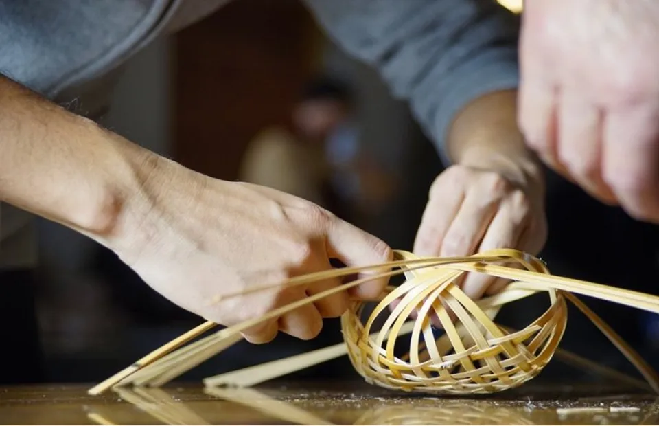 장인의 손을 찍은 클로즈업. 가늘게 쪼갠 대나무를 사용하여 둥근 바구니를 짜고 있는 모습. 대나무 섬유가 교차하며 섬세한 전통 공예품이 만들어지는 과정이 보여진다.