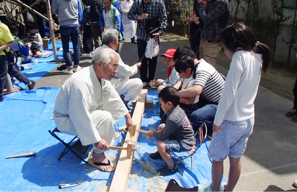 야외 푸른 시트 위에서 연배의 장인이 아이들에게 목공을 가르치고 있는 풍경. 장인과 소년이 마주 보고 긴 목재에 나무망치를 사용하고 있다. 주변에는 이를 지켜보는 어른들과 아이들이 모여 있다.