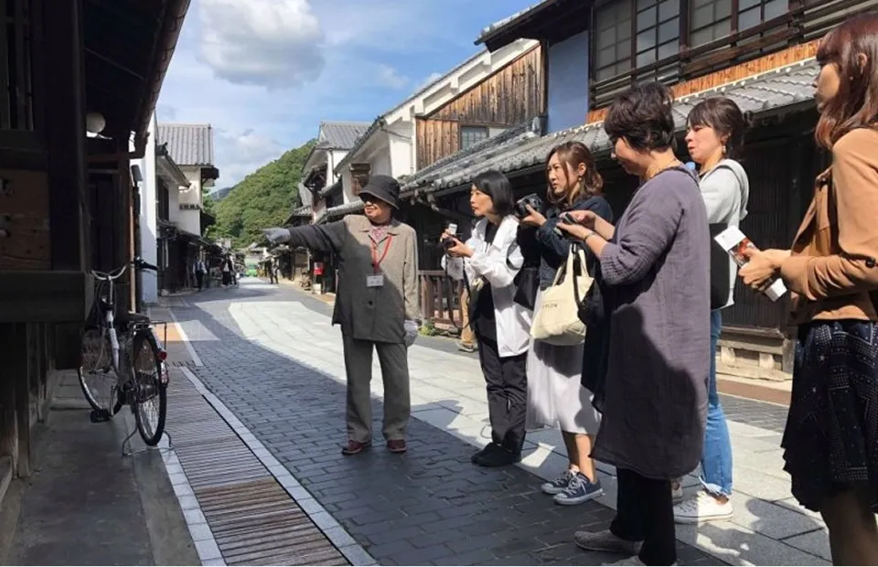 전통 가옥이 늘어선 돌담길에서 여성 가이드가 관광객에게 해설을 하고 있는 풍경. 모자를 쓴 가이드가 왼쪽을 가리키고, 몇 명의 여성 고객이 열성적으로 이야기를 듣거나 카메라를 향하고 있다. 역사가 느껴지는 조용한 마을 산책 모습.