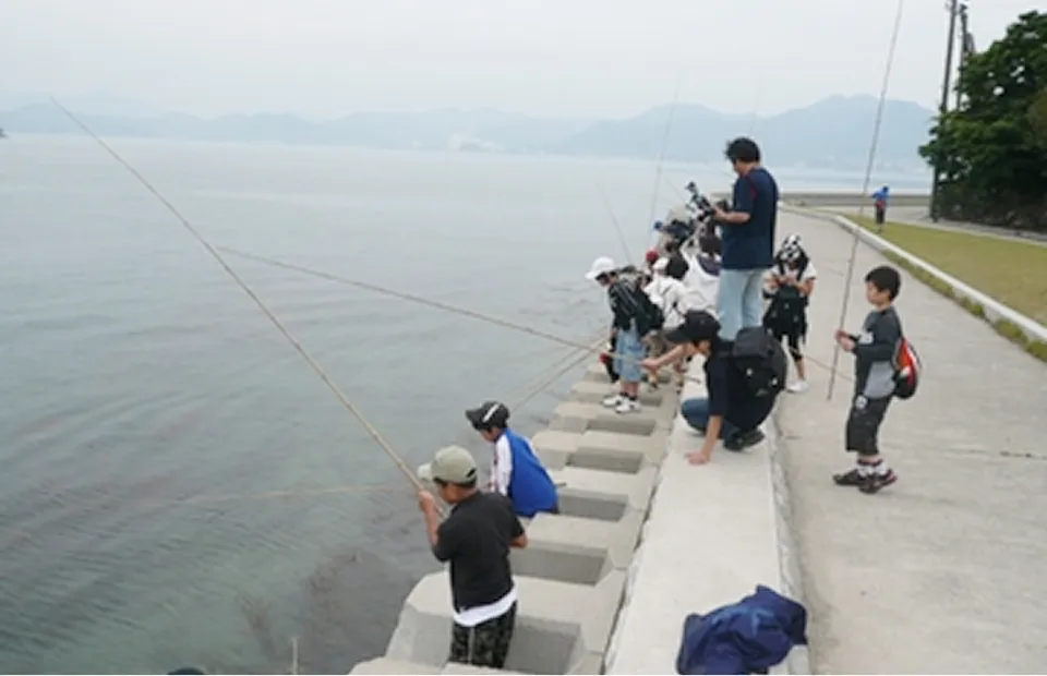 흐린 하늘 아래 해안가 제방에 일렬로 늘어서서 낚시를 즐기는 아이들과 어른들의 모습. 많은 사람이 긴 낚싯대를 해수면으로 향하고 조용히 물고기가 잡히기를 기다리고 있다. 멀리에는 안개 낀 산맥이 펼쳐지는 평온한 체험 풍경.