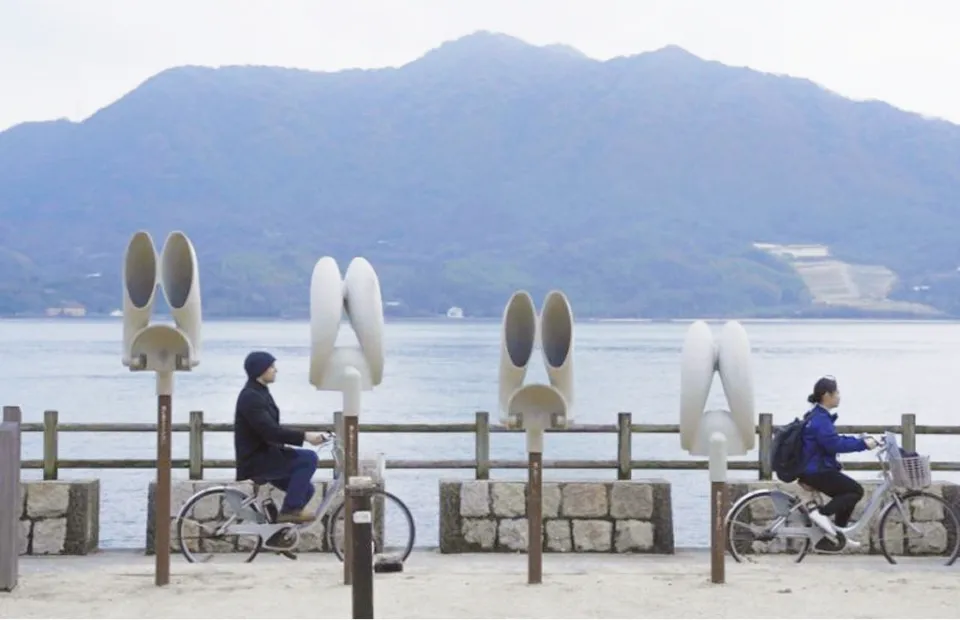 해안 도로를 자전거로 달리는 남녀의 풍경. 도로변에는 귀 모양이나 원통 모양의 하얀 집음 오브제가 늘어서 있다. 배경에는 온화한 바다와 화면 가득 펼쳐진 큰 산이 보인다.