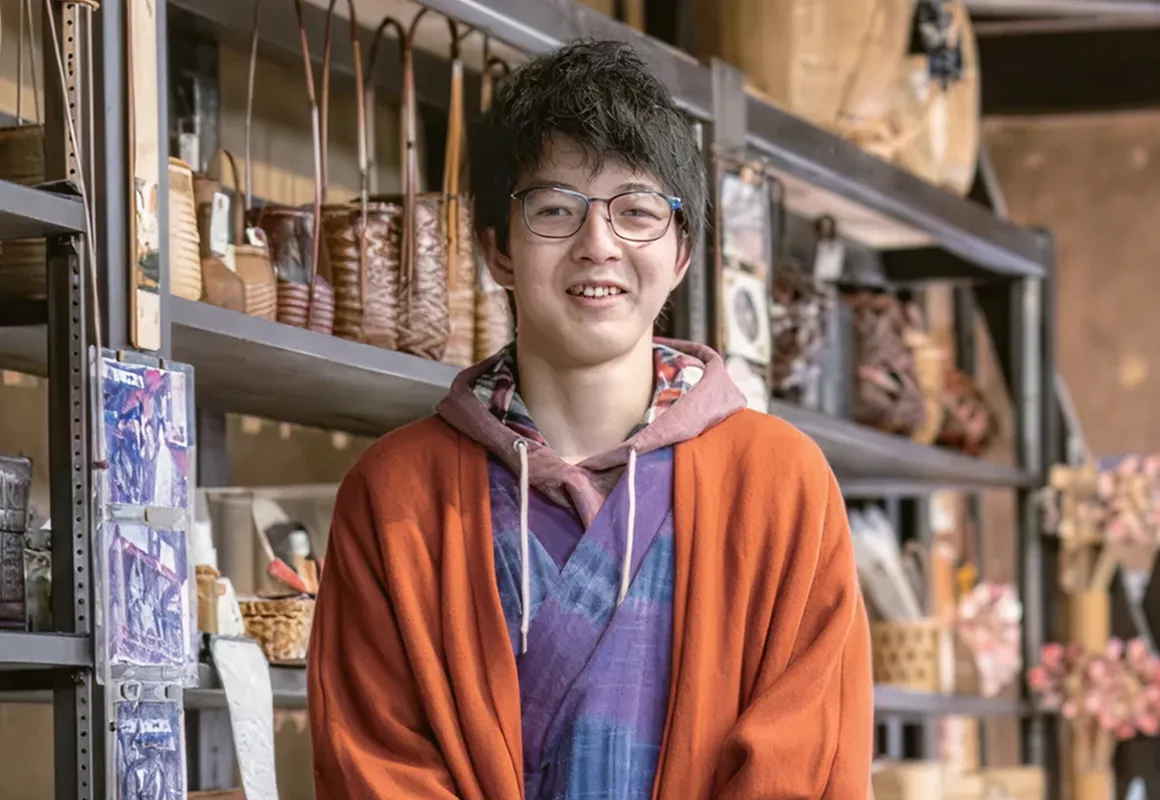 站在陳列竹編籃子的架子前、戴眼鏡的年輕男性。身穿紫色如和服般的服裝，外搭橘色羽織，呈現傳統與現代交融的造型。面帶溫和笑容，讓人感受到他是投入傳統文化的新一代傳承者。