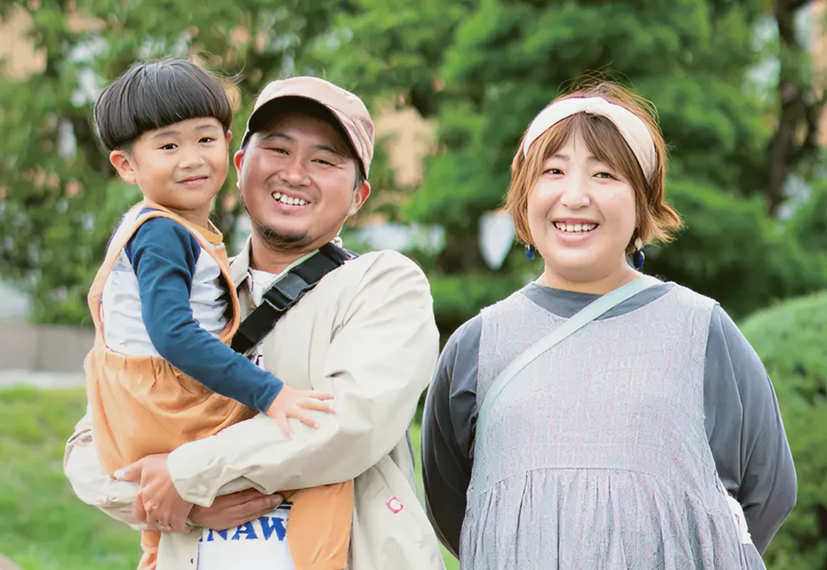 在綠意盎然的戶外，露出燦爛笑容的年輕夫妻與年幼男孩組成的三口之家。父親抱著男孩，母親在旁微笑。是一張象徵移居後幸福充實生活的溫暖家庭照。