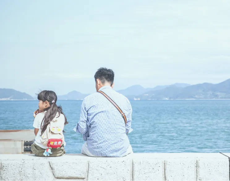 以蔚藍海面為背景，坐在防波堤上交談的親子（父親與年幼女兒）背影。女孩背著背包，兩人凝望平靜的地平線。彷彿流淌著家人安靜而溫暖時光的畫面。