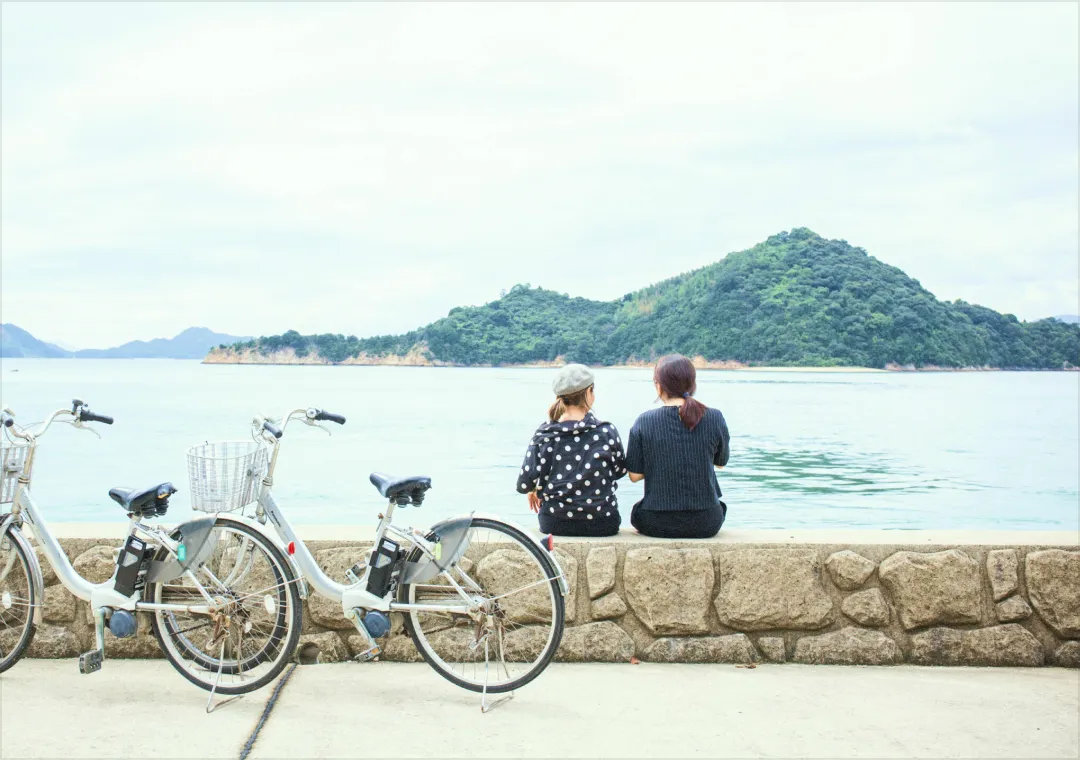 兩人坐在海邊石階上，眺望著浮著小島海景的背影，前方停著兩台疑似租借的自行車。