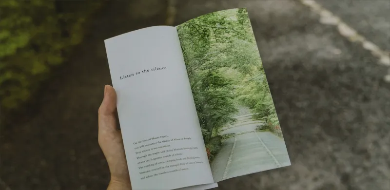 雙手拿著翻開的手冊，背景為森林中的小徑